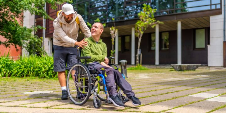 A man with special needs who is in a wheelchair, smiling together with another man who is his carer.